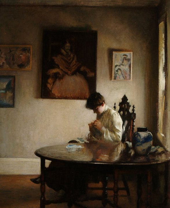 Woman seated at a large round table. She is looking down at her hands where she works on something. Behind her are large portraits. The light is dramatic and comes from the right side where there is a window. 