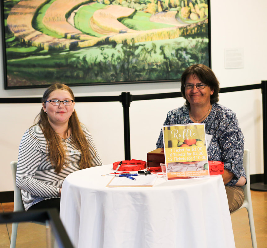 Picture of a young white woman with glasses and long hair, and an older woman with short brown hair sit behind a table selling raffle tickets