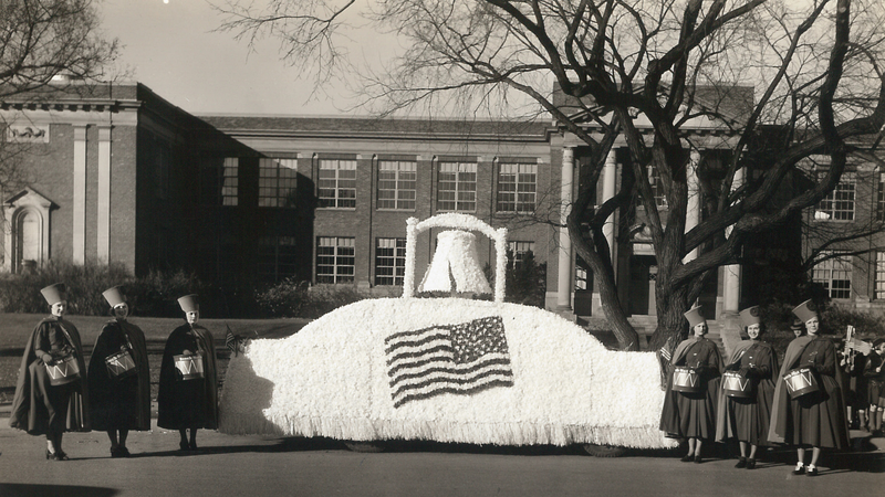 Six sample girls flank a large float. The float is white with an American Flag on the front and the Liberty Bell on top. The float and sample girls stand in front of the Canajoharie High School on Erie Blvd. 
