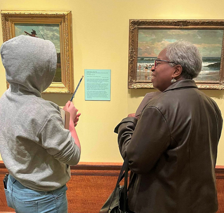 Picture of A person in a gray hoddie and a Black woman in a leather coat look at two paintings by Winslow Homer.