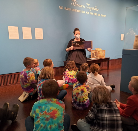 Picture of a white woman sitting in a museum gallery speaking and reading a book to a group of young children. 