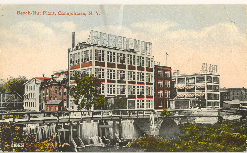 Postcard of the Beech-Nut Factory. It is a drawing but with color. There are several large buildings with many windows. In front of the building water comes down almost like a waterfall. 
