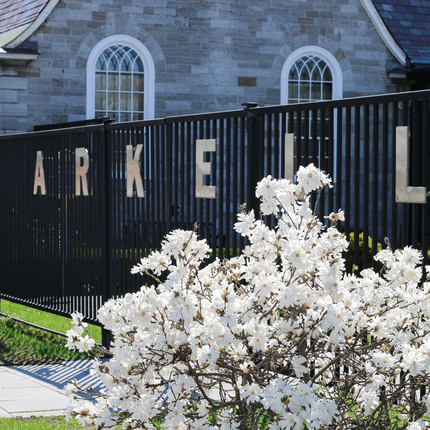 Picture of a white flowering magnolia bush with a black iron fence behind with the words: ARKELL spelled out. Behind is the side of a building made out of blocks of gray stone. 